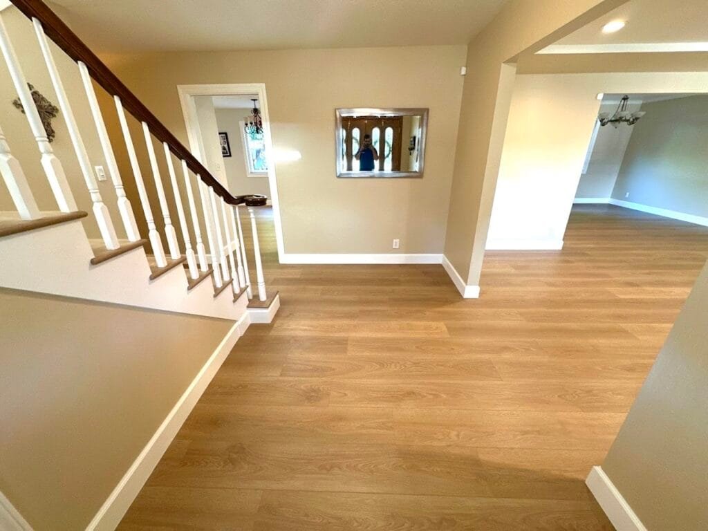 A hallway with light wood floors, beige walls, a staircase with white spindles, and a mirror on the wall showcases the quality work of a construction company specializing in kitchen remodels. Two adjoining rooms are also visible.