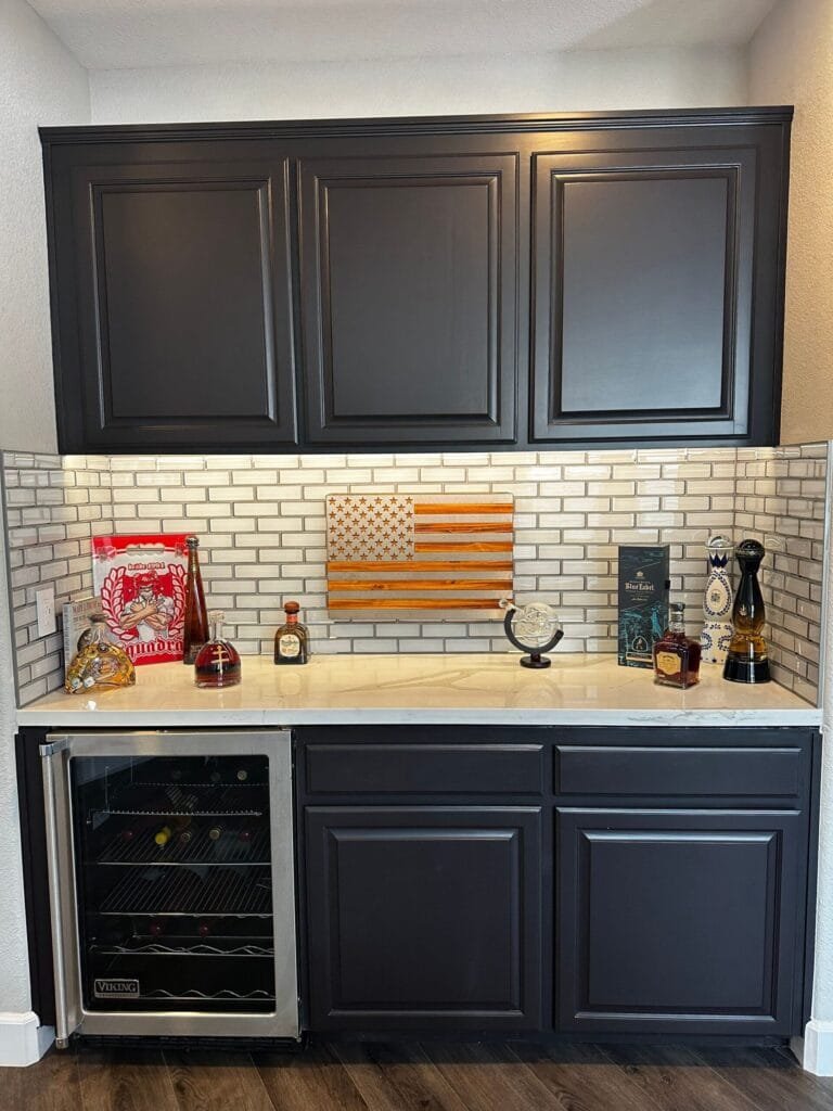 Kitchen Remodeling Services in Blackhawk, CA 3 A home bar area with dark cabinets, a mini fridge, bottles, glassware, books, and an American flag wall art above a white countertop with a subway tile backsplash—perfect inspiration for your next kitchen remodel.