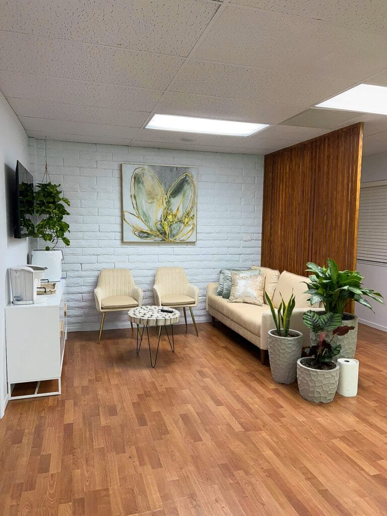 Modern waiting room with a beige couch, two chairs, potted plants, a wall painting, and wooden flooring—perfect inspiration for your next home remodel. A partition with vertical wood slats elegantly separates the area.