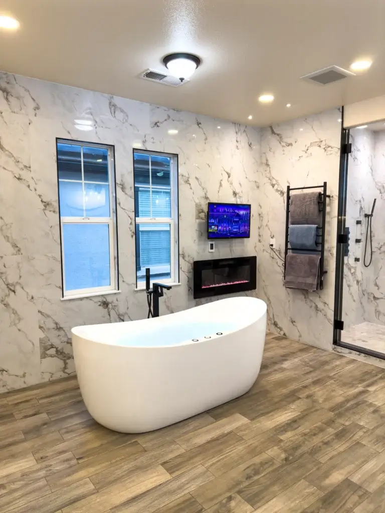 Modern bathroom by a leading construction company, featuring a freestanding white tub, marble walls, wood-look flooring, wall-mounted TV above an electric fireplace, towel rack, and glass shower enclosure—a showcase bath remodel.