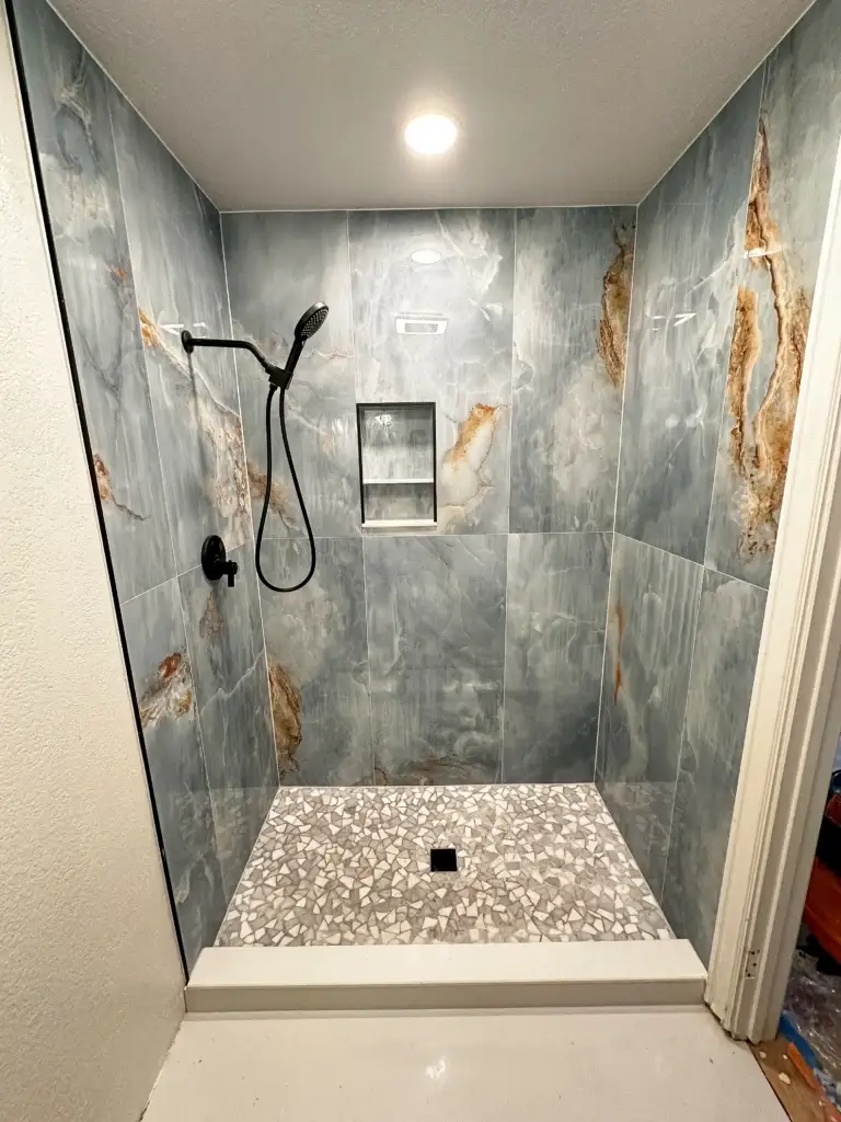 Modern walk-in shower with large marble-patterned tiles, a handheld showerhead, built-in shelves, and a stone-patterned floor under recessed ceiling lighting—perfect for your next bath remodel by an expert construction company.