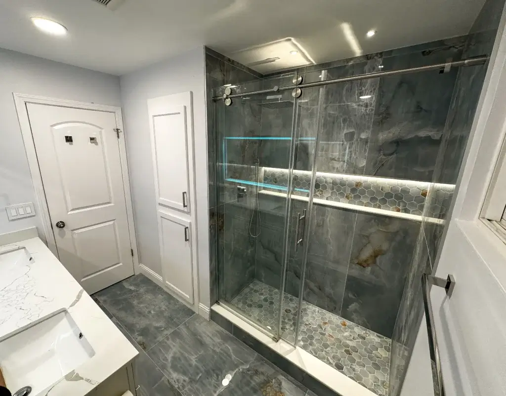Modern bathroom with marble tile, a glass-enclosed shower featuring built-in shelves and accent lighting, a white vanity, and two closed white doors—perfect inspiration for your next bath remodel or home remodel by a trusted construction company.