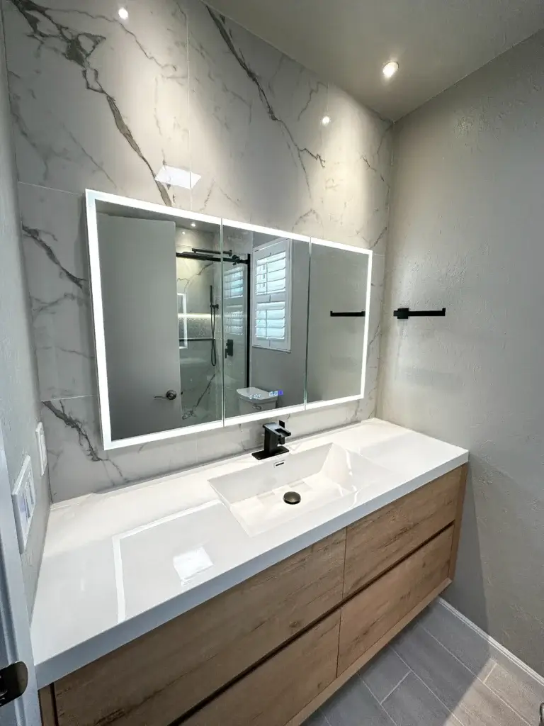 Modern bathroom with a large backlit mirror, marble accent wall, minimalist sink, black faucet, light wood vanity, and neutral tile flooring—perfect for a sophisticated bath remodel or stylish home renovation.