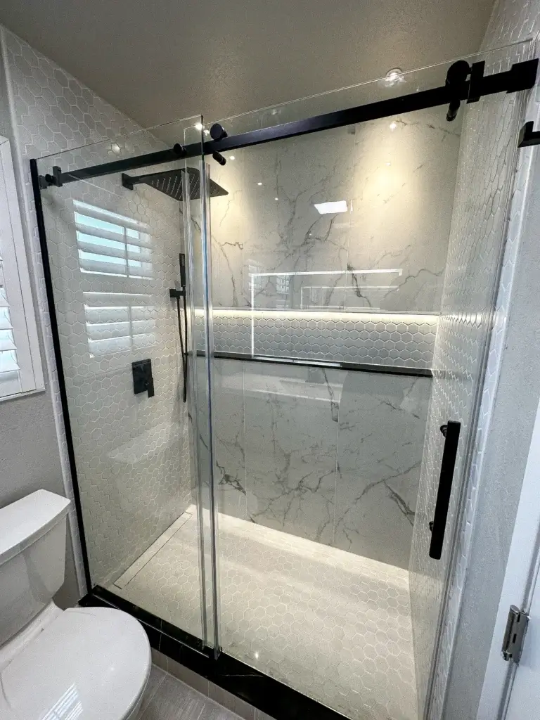 Modern walk-in shower with glass sliding doors, black hardware, marble-patterned walls, hexagon tile accents, and built-in shelf with LED lighting—perfect for your next home renovation or home remodel. Toilet visible to the left.