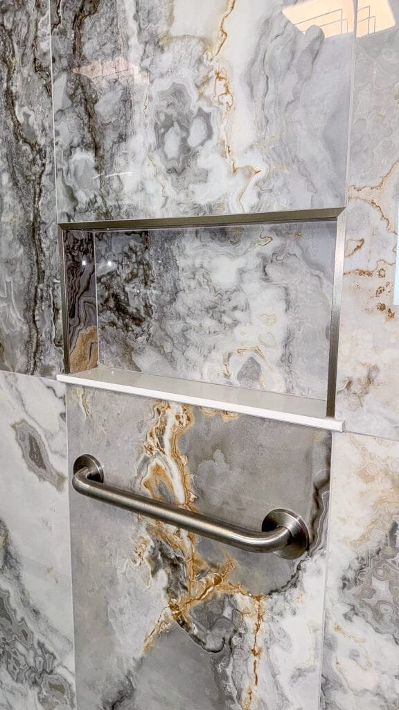 Elevate your home renovation with this shower wall featuring gray and gold marble-patterned tiles, a rectangular recessed shelf with a metal frame, and a stainless steel grab bar below the shelf.