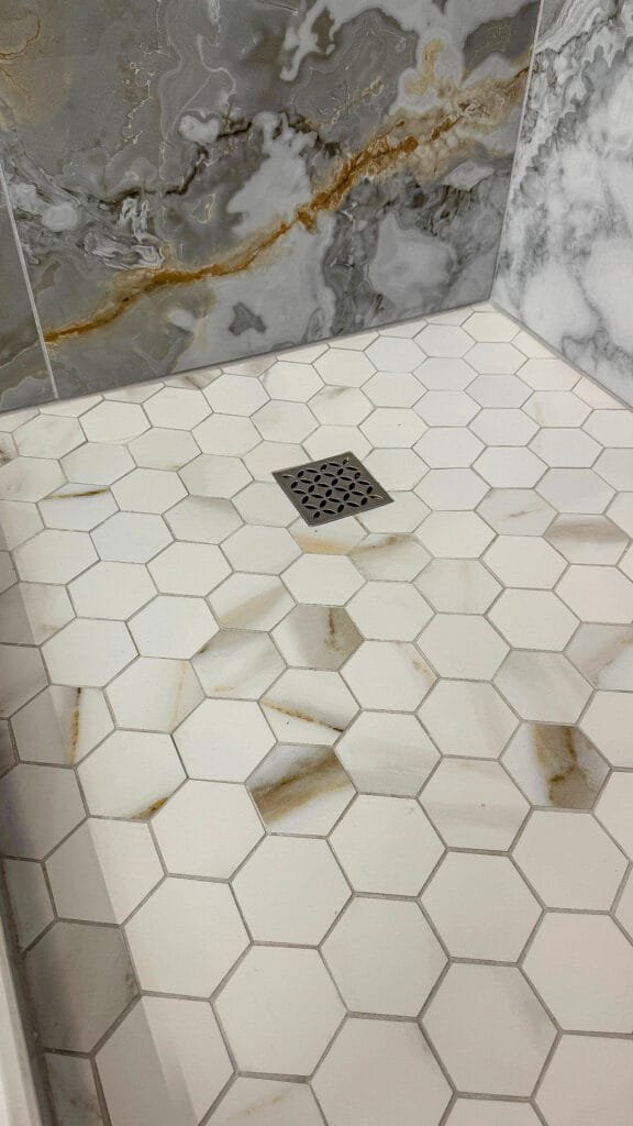 A shower floor with white hexagonal tiles featuring brownish stains, a square metal drain in the center, and gray marbled wall tiles in the background—ideal for a bath remodel by a trusted construction company.